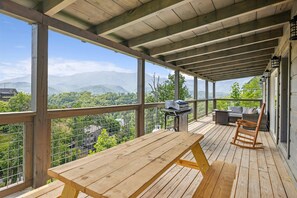 Una terraza de madera con mesa de picnic y parrilla, con vistas a un paisaje montañoso.