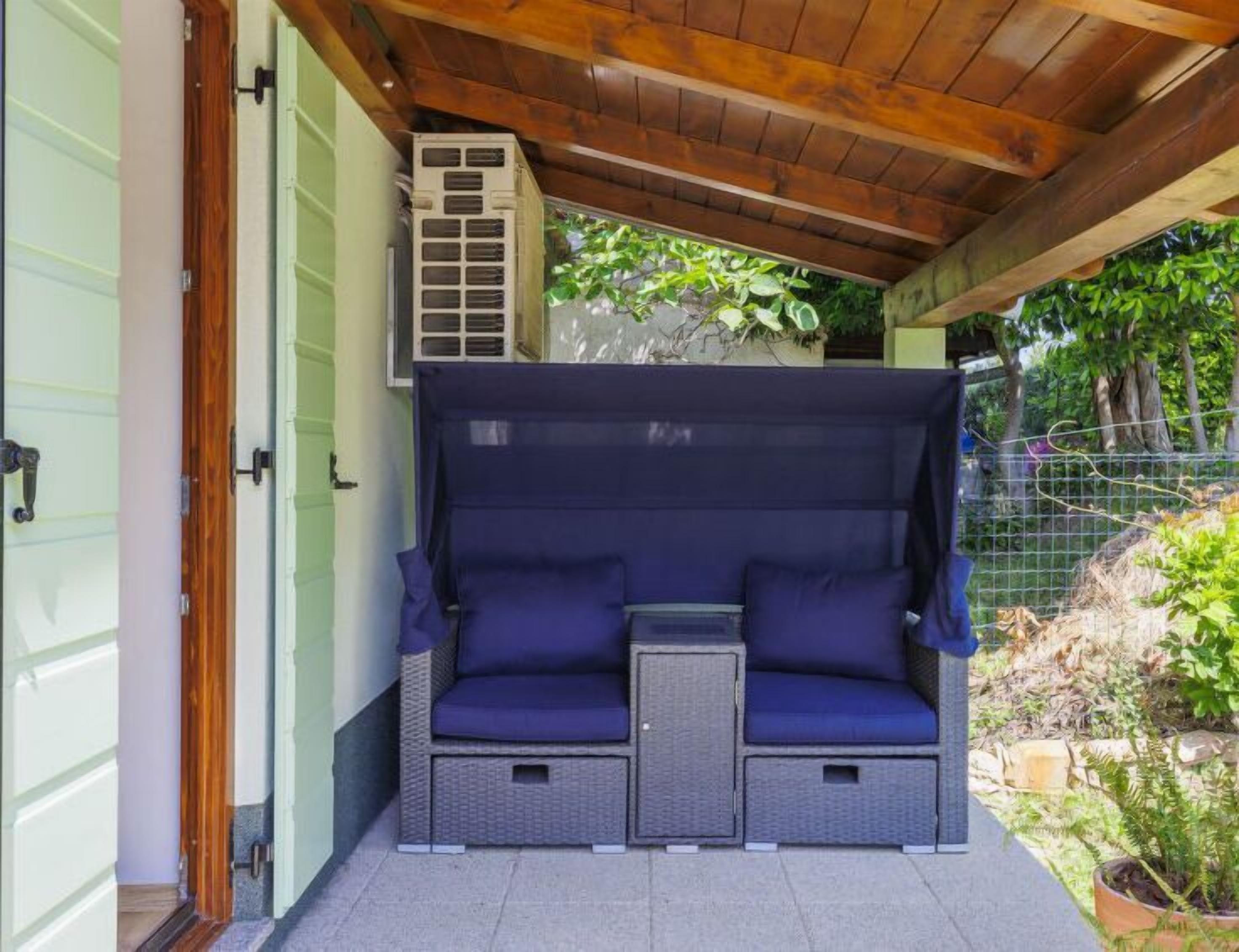 A covered outdoor seating area with two cushioned benches and a small table, adjacent to a building with a green door and an air conditioning unit.
