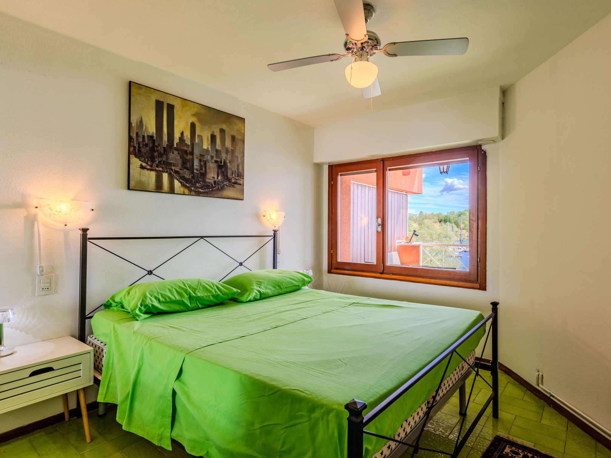 Een slaapkamer met een bed, nachtkastje, schilderij, plafondventilator en een raam met uitzicht.