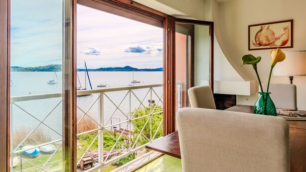 A room with a balcony overlooking a lake with boats, a chair, a table, and a vase with flowers.