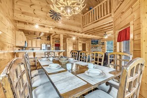 Interior de una cabaña de madera con una mesa de comedor lista para seis comensales, paredes de madera y una araña.