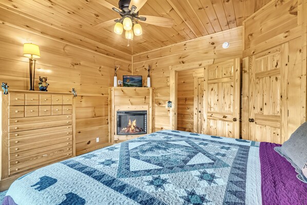 Una acogedora habitación tipo cabaña de madera con chimenea, paredes de madera, una cama con colcha, un aparador y un ventilador de techo.