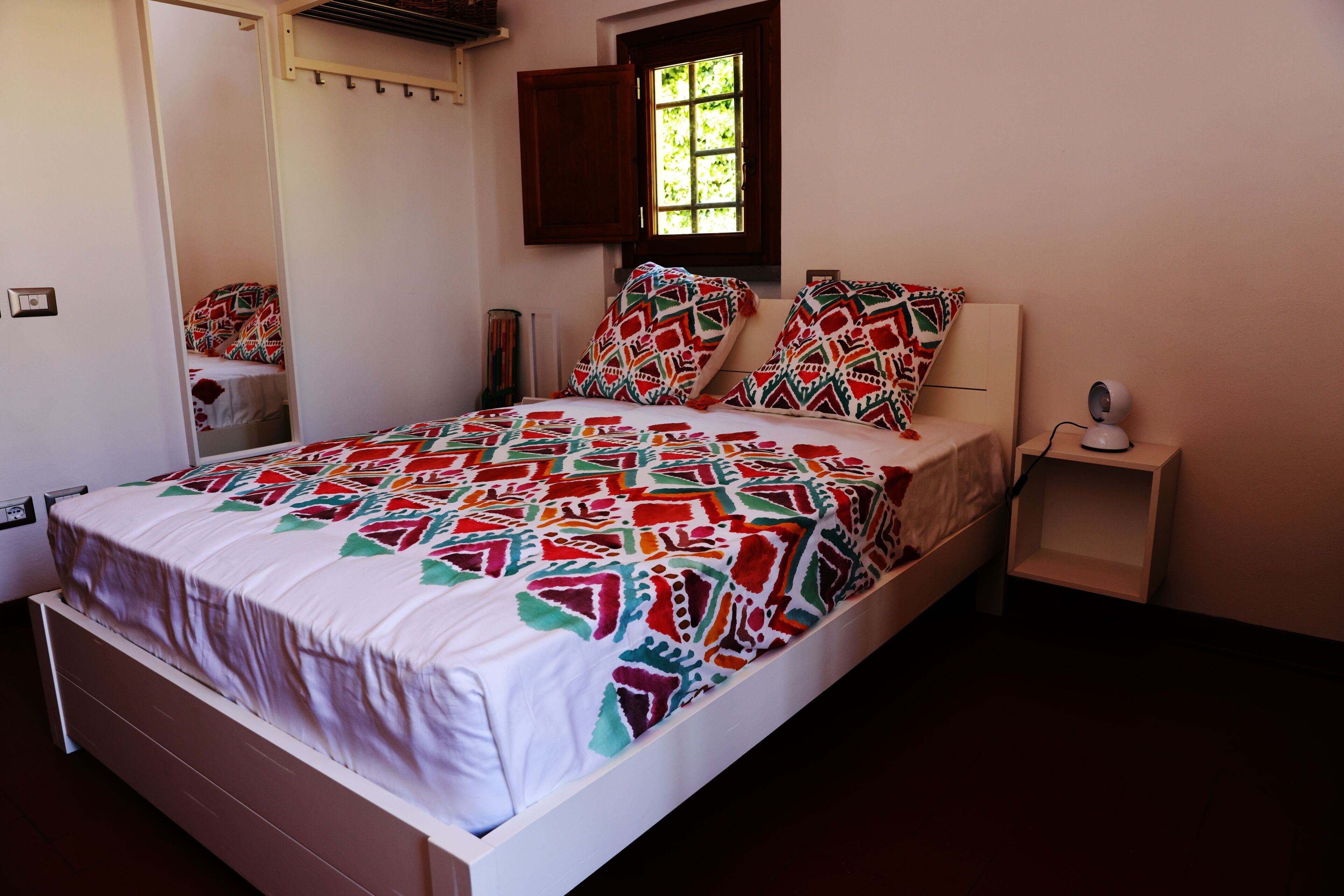 Een slaapkamer met een bed, een raam, een nachtkastje en een spiegel.