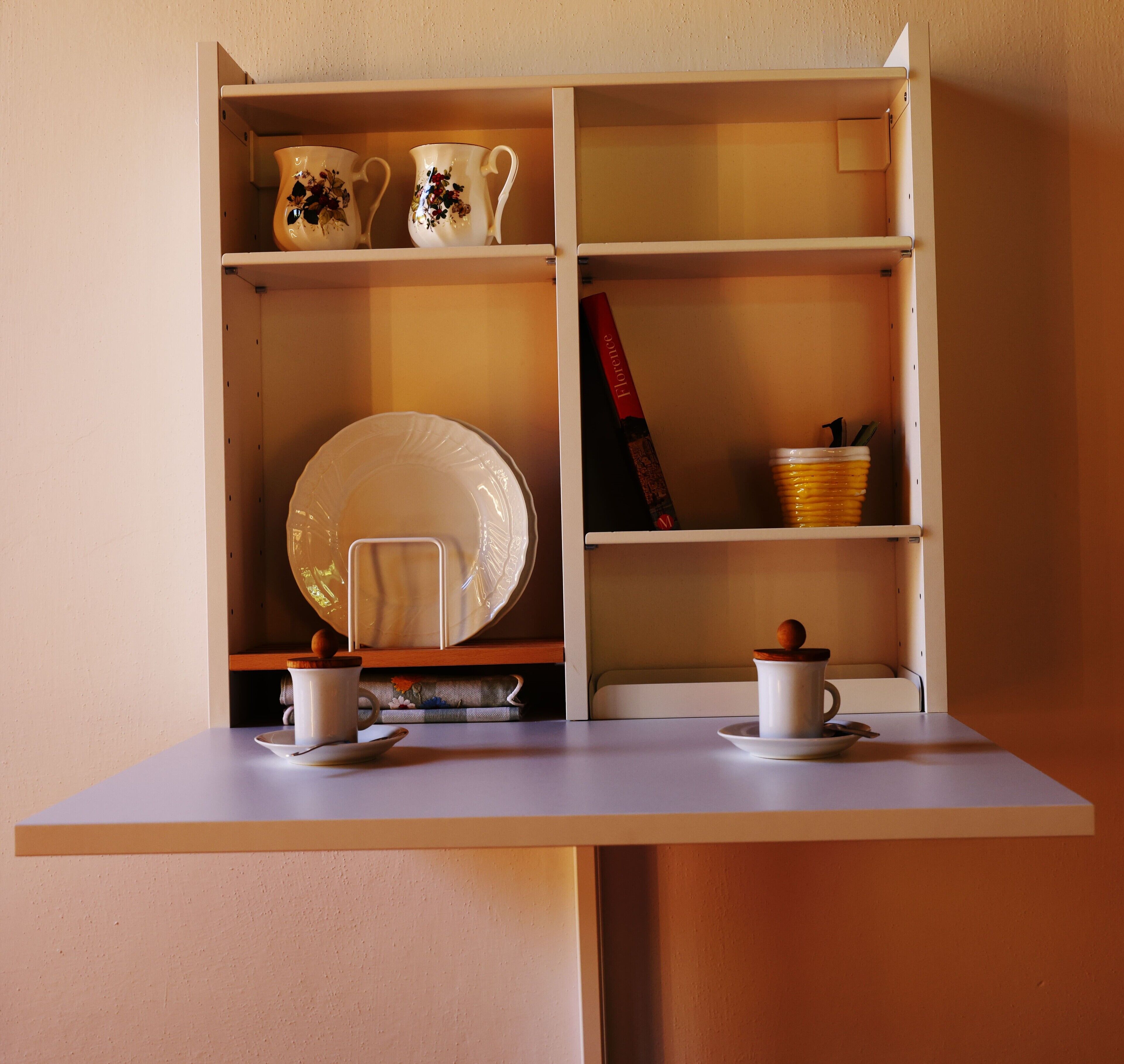 A white cabinet with two shelves, each holding a white teacup and saucer, a white plate, and a red book titled "Florence".
