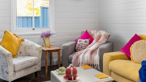 A cozy living room with a white sofa, a yellow armchair, and a gray armchair. There is a small wooden table with a vase of flowers and a decorative item. A window with blue shutters is visible in the background. - Olive Nook in Katoomba (Katoomba)