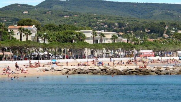 Une plage où l’on peut voir des gens se prélasser au soleil et nager, une digue rocheuse et des bâtiments en arrière-plan.