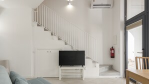 Een moderne woonkamer met een trap, een flatscreen televisie op een witte standaard, een bank en een eettafel.