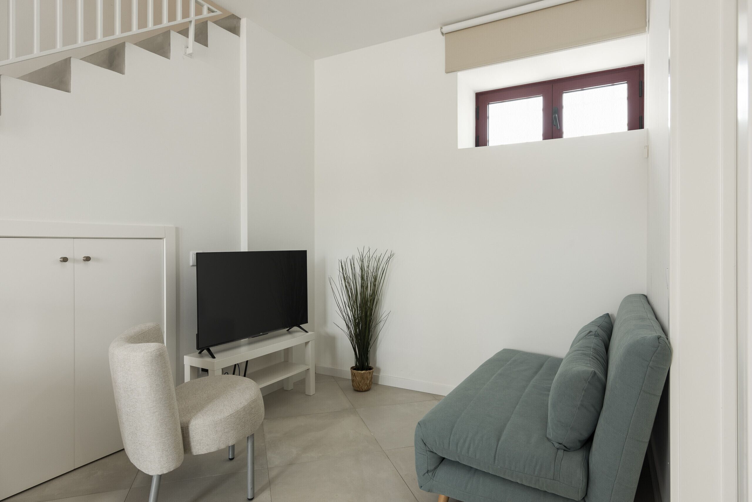 A modern living room with a flat-screen TV, a grey sofa, a white chair, a small potted plant, and a window with a blind.