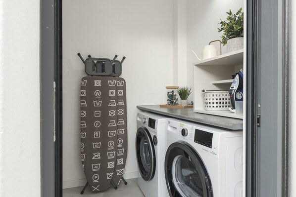 A laundry area with two front-loading washing machines, a drying rack with various symbols, and a shelf with a plant and a basket.