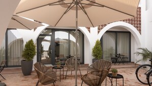 Een terras buiten met rieten stoelen, een grote parasol en potplanten.