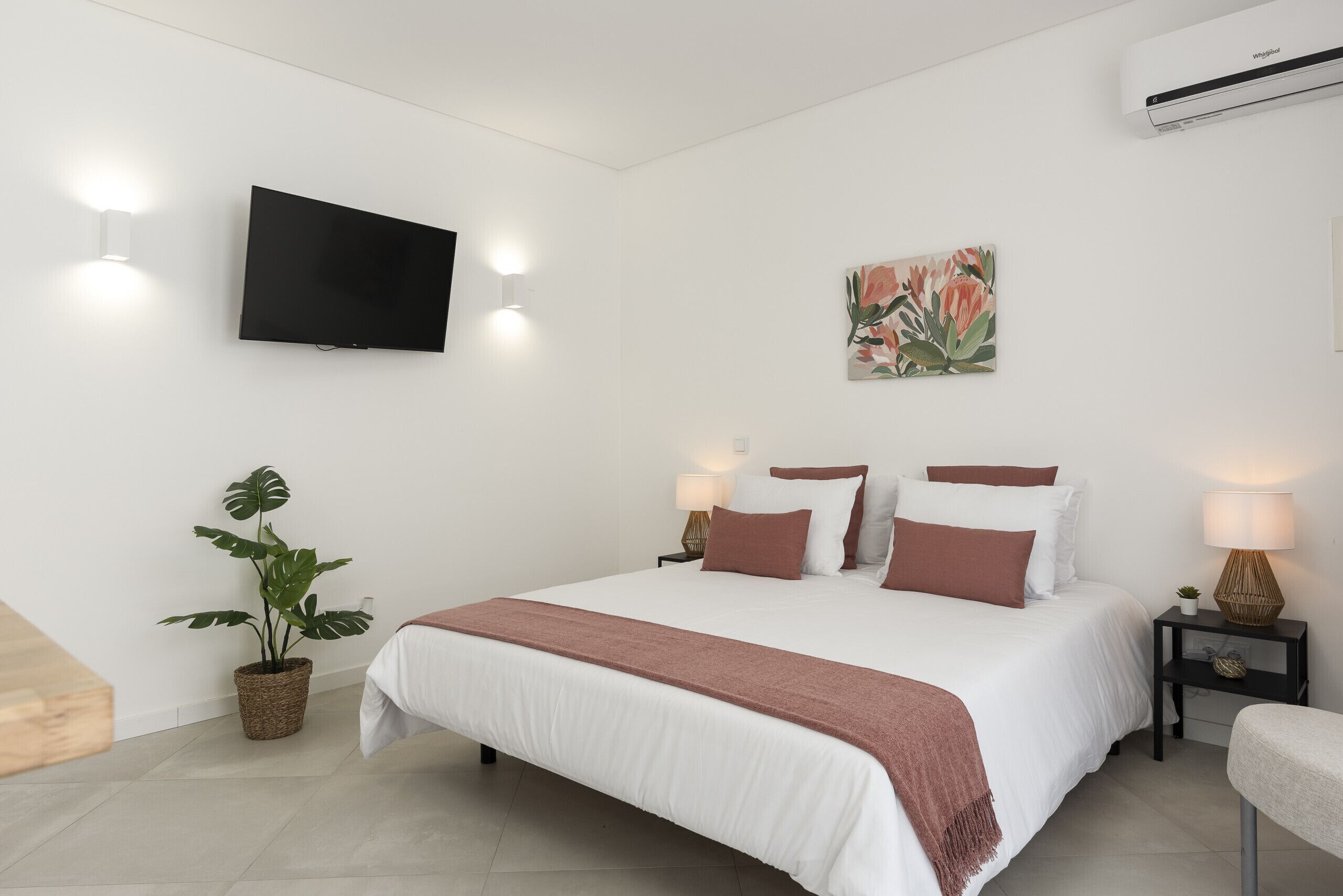 A modern bedroom with a large bed, a flat-screen TV, a potted plant, and a wall-mounted air conditioner.