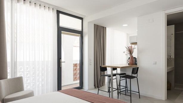 Moderni hotellihuone, jossa on sänky, työpöytä kahdella tuolilla, ruokailutila ja näkymä ulos.