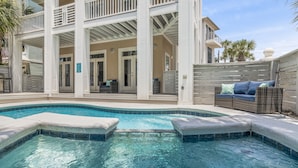 Una piscina con un porche cubierto, un edificio blanco con varias puertas y mobiliario exterior.