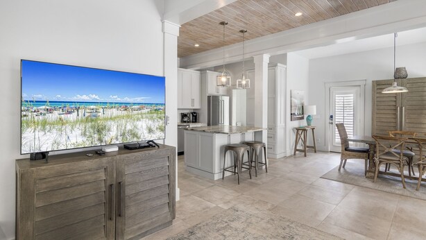 Una cocina amplia con una isla grande, un comedor con mesa y sillas de madera, y un televisor mostrando una escena de playa.
