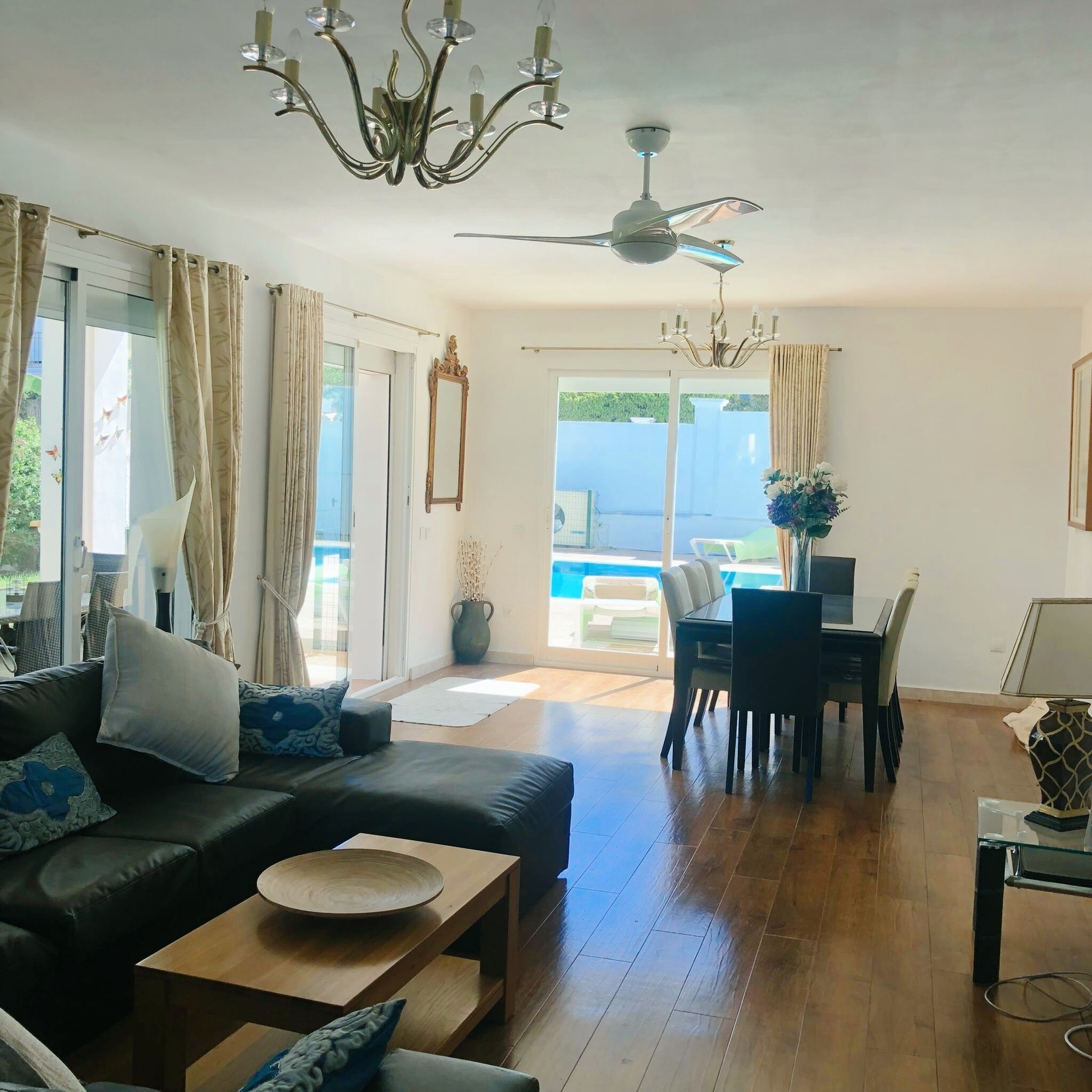 Un vaste salon avec un canapé noir, une table basse en bois et un coin repas comprenant une table et des chaises. Un lustre est suspendu au plafond, et un ventilateur de plafond est visible.