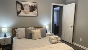 Un lit bien fait, avec une couette blanche et plusieurs oreillers, une table de chevet avec une lampe, et un tableau abstrait accroché au mur.