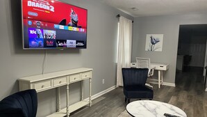 Un salon avec un téléviseur à écran plat fixé au mur, qui diffuse différents services de streaming. On y trouve une console blanche, deux fauteuils sombres et une table basse en marbre sur laquelle repose une télécommande.