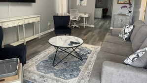 Un salon comprenant un téléviseur à écran plat, un fauteuil bleu foncé, un canapé gris, une petite table basse ronde et un tapis à motifs.
