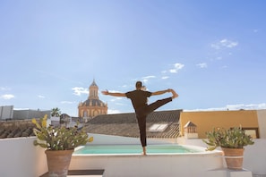 En person udfører en yogastilling på et tag med en pool og potteplanter med kaktusser, med udsigt over en bygning med et markant tårn.