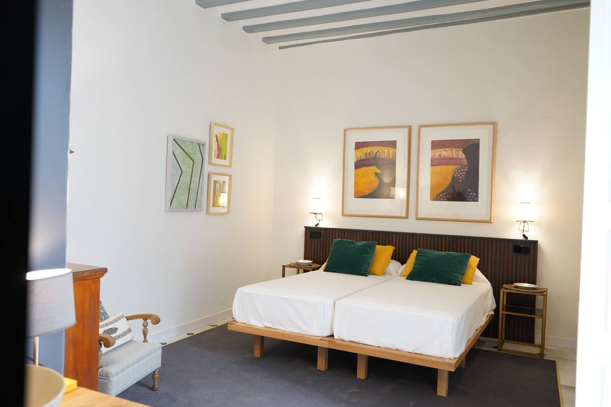 Uma cama de casal com estrutura de madeira, duas mesas de cabeceira e um pequeno espaço de estar com uma poltrona e uma mesa.