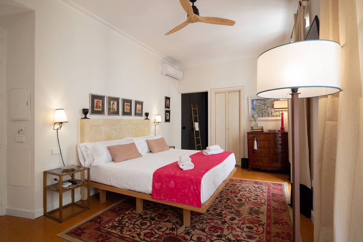 Quarto com cama, mesas de cabeceira, ventoinha no teto, cômoda e uma janela grande com cortinas.
