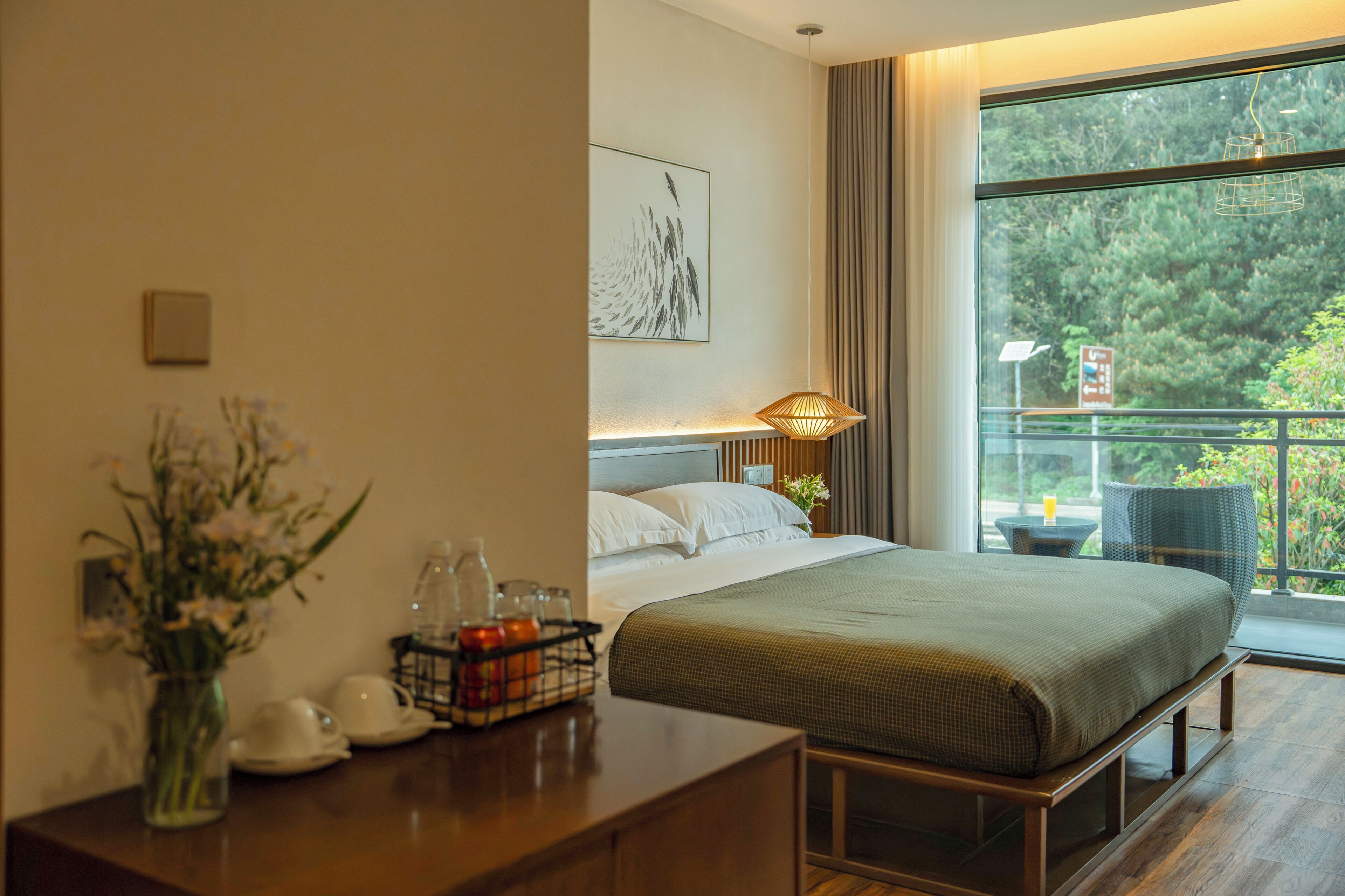 Une chambre d’hôtel avec un lit, un bureau agrémenté d’un vase de fleurs, une fenêtre donnant sur des arbres et une lampe.