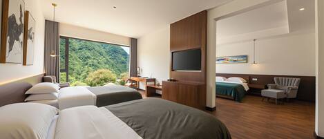 A hotel room with two beds, a desk, a TV, and a view of a forest.