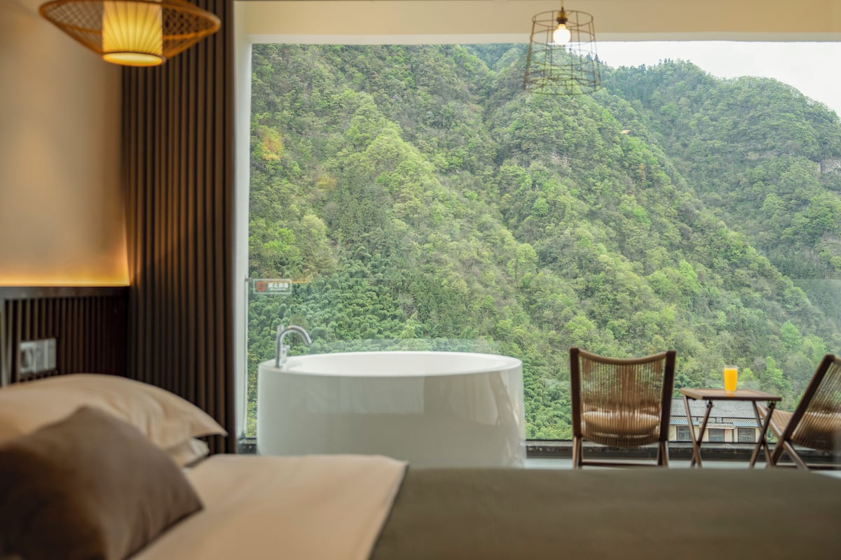 Une chambre avec une grande fenêtre, une baignoire, deux fauteuils en osier et une petite table sur laquelle se trouve un verre de jus d’orange.