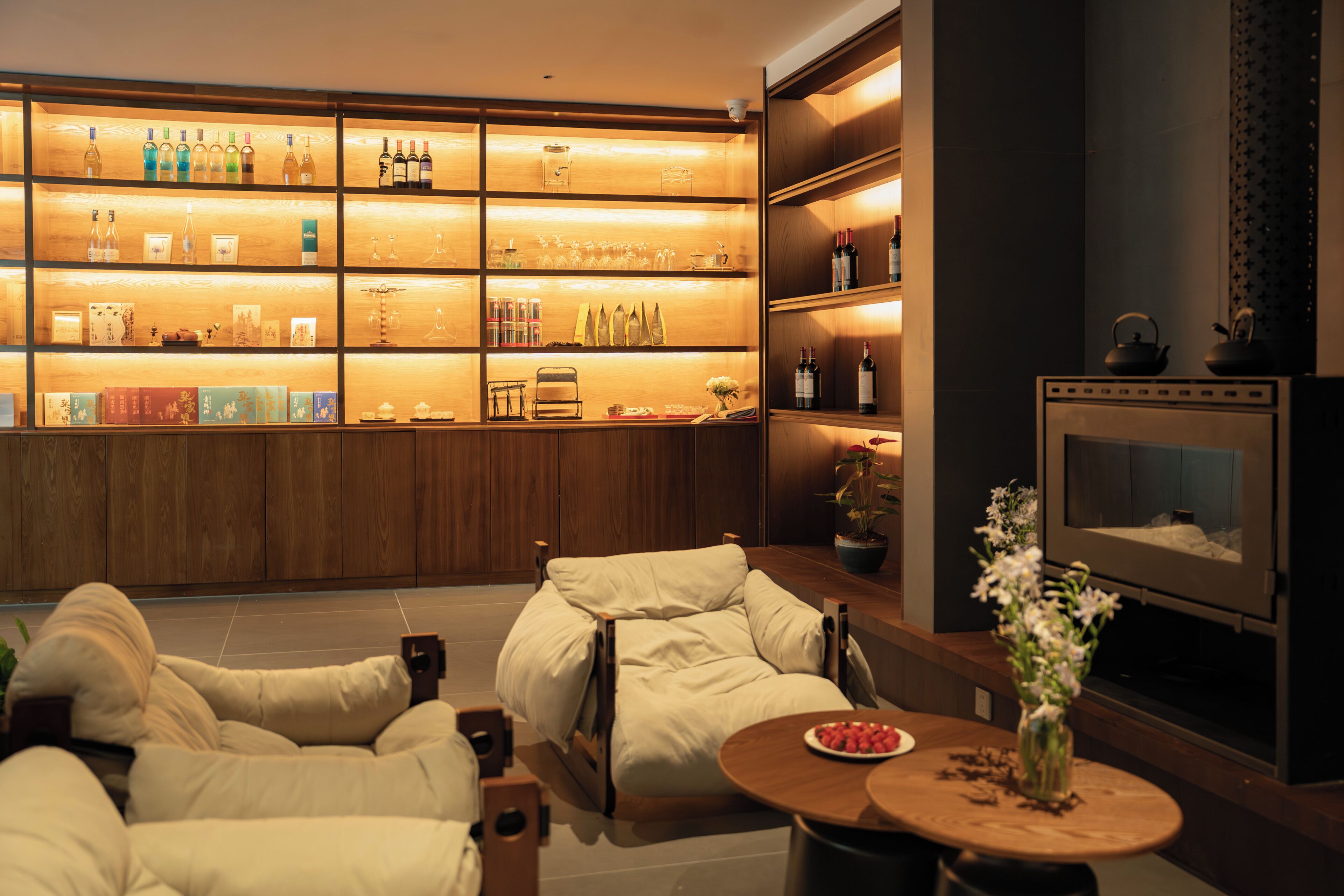 A modern living room with a sofa, a round table with a vase, and a TV. There are shelves with various items and a wooden cabinet in the background.