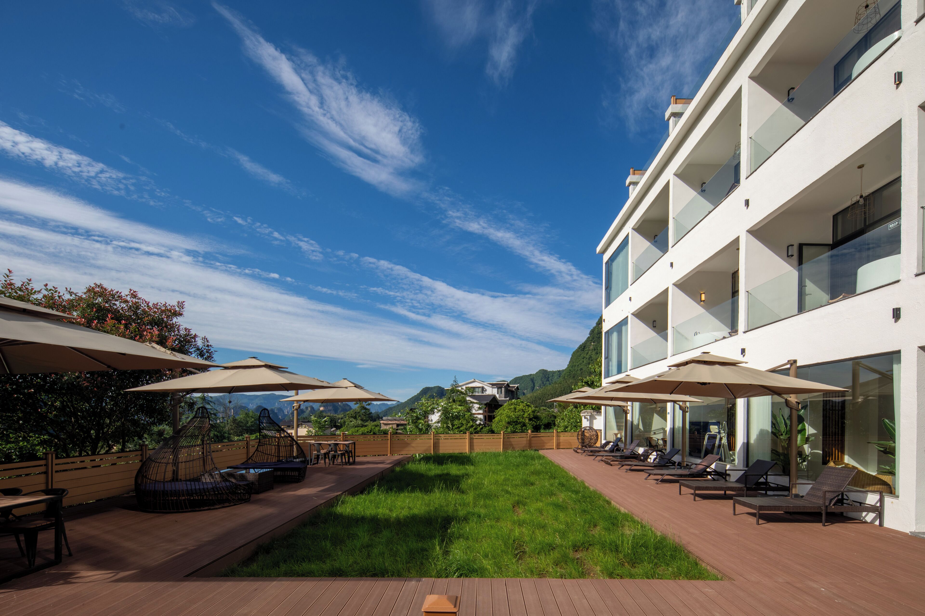 A modern outdoor patio with lounge chairs and umbrellas, a wooden deck, and a view of a mountain.