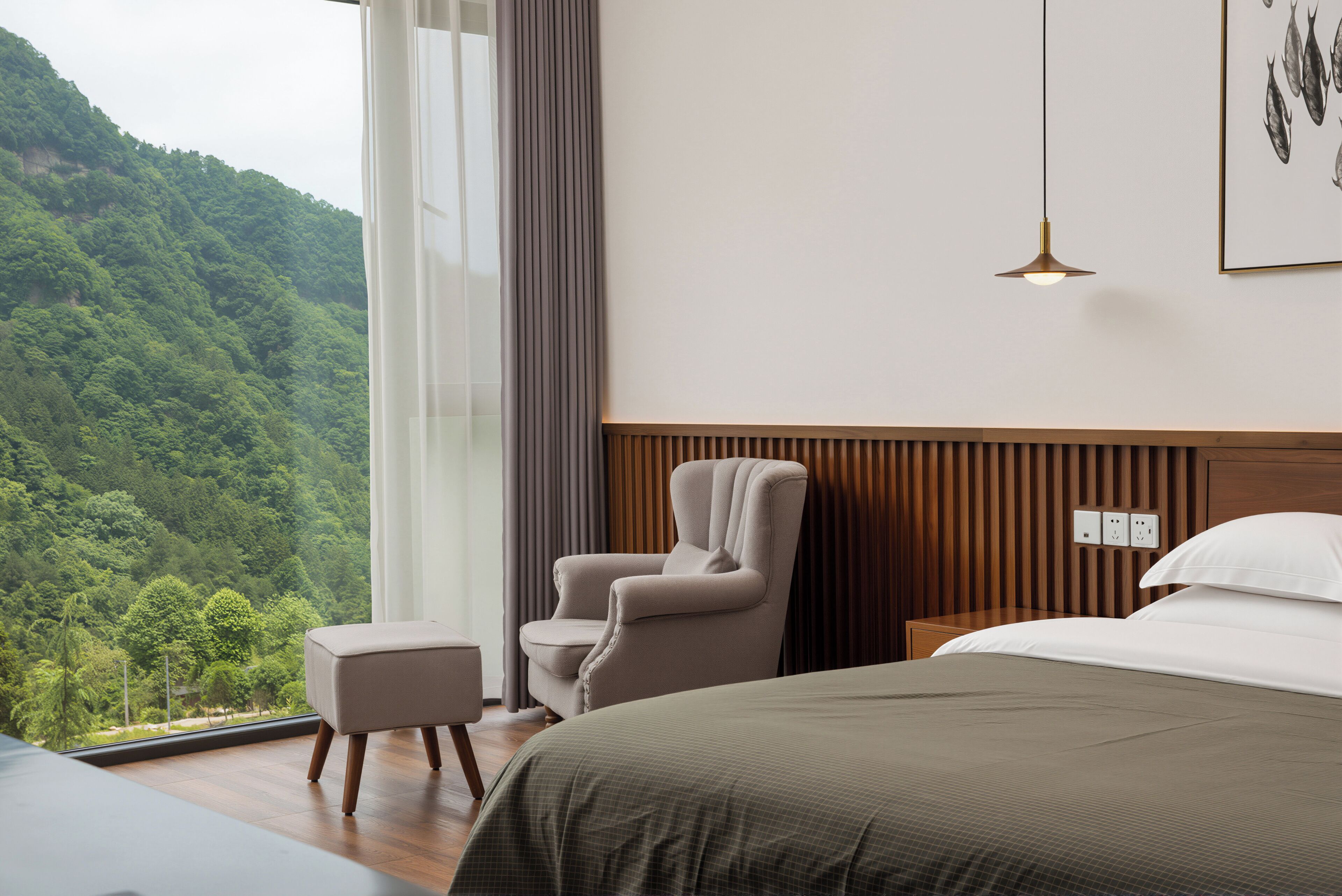 Une chambre à coucher avec un grand lit, une chaise, une table de chevet, une commode, une lampe suspendue et une fenêtre donnant sur une zone boisée.