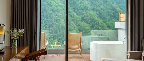 A modern hotel room with a large sliding glass door leading to a balcony with a view of a forested area.