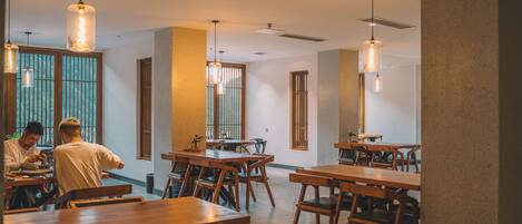 A spacious dining area with wooden tables and chairs, pendant lighting, and large windows with vertical blinds.