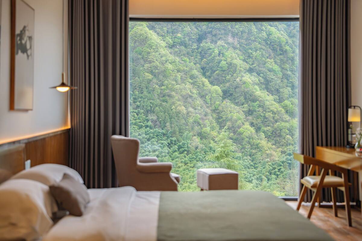 Une chambre d’hôtel avec un lit, un bureau, une chaise et une vue sur une forêt, visible à travers une grande fenêtre.