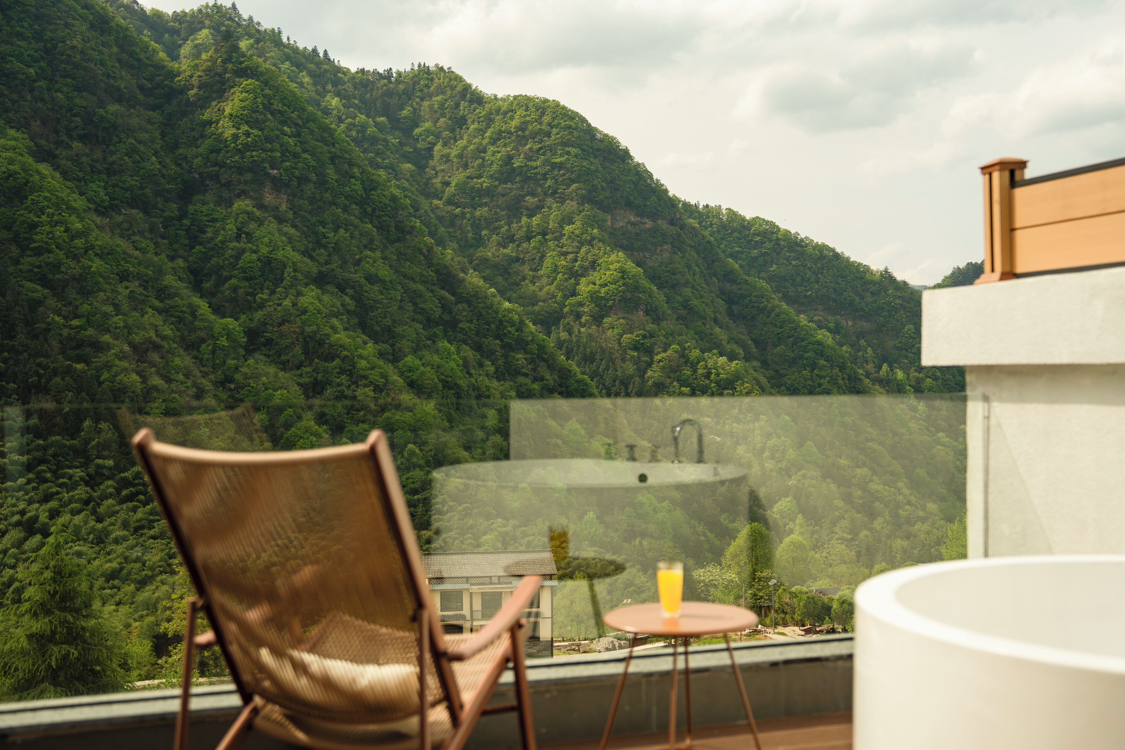 Un balcone con una sedia, un tavolino con un bicchiere di succo e vista su una montagna verde e rigogliosa.