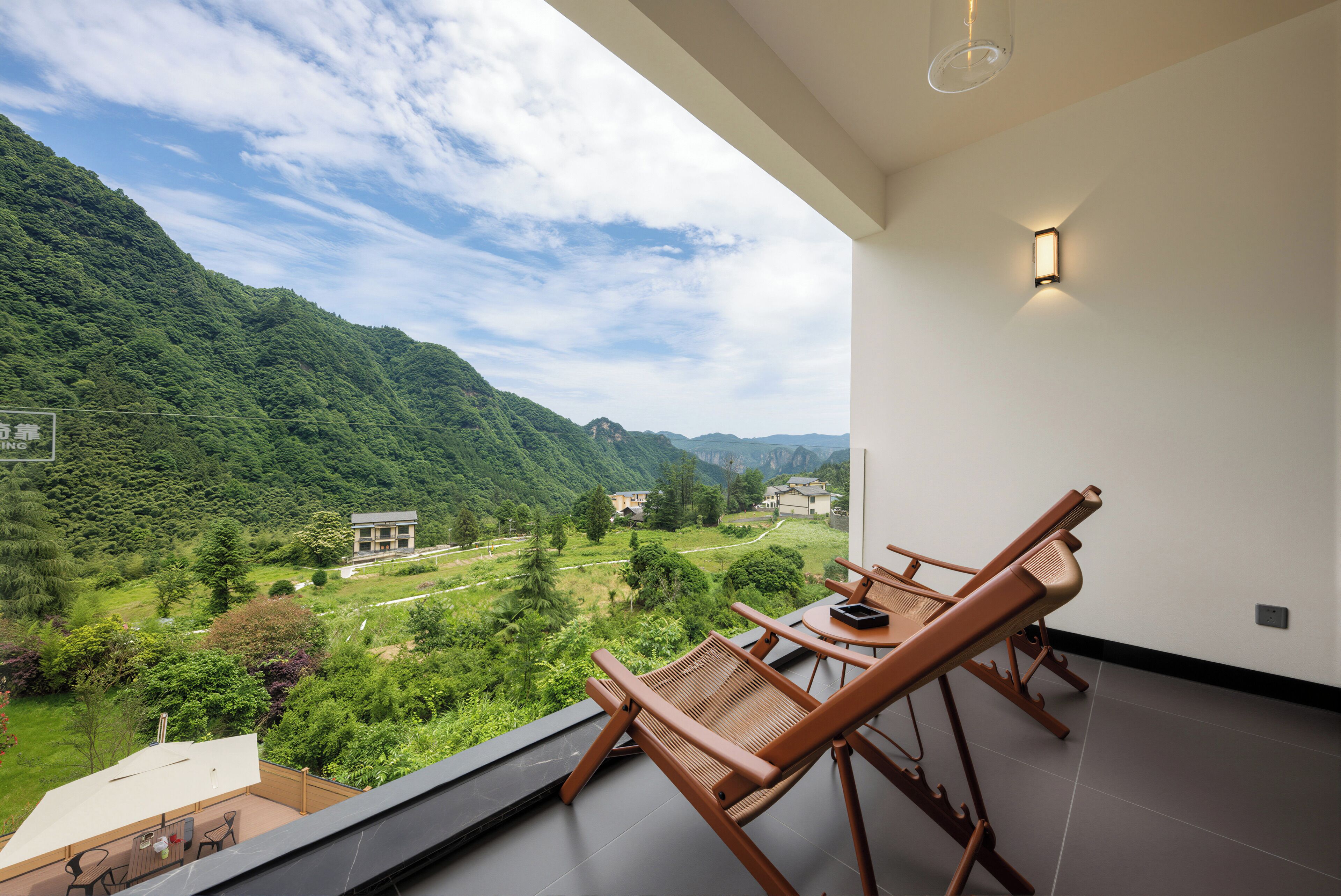 A balcony with wooden chairs and a table, overlooking a lush valley and mountains.