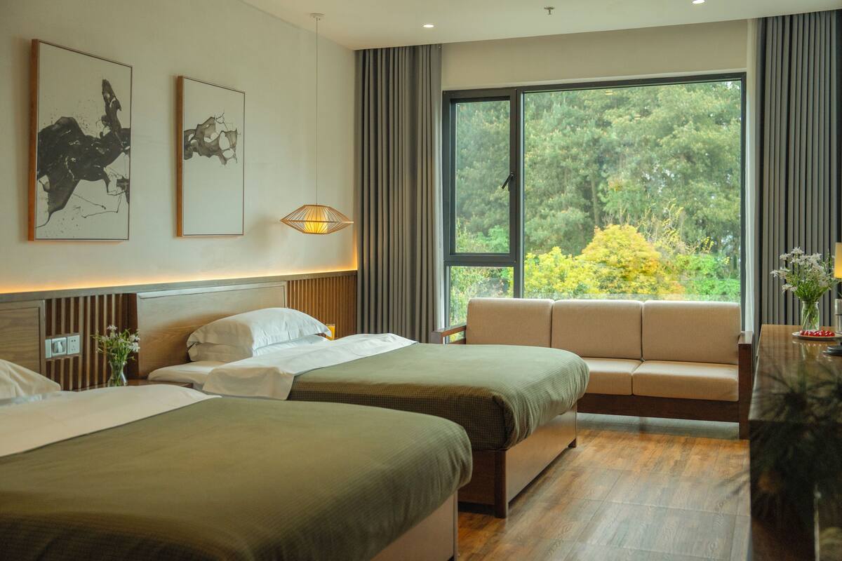 Une chambre d’hôtel avec deux lits, un canapé, une grande fenêtre donnant sur des arbres et des œuvres d’art accrochées au mur.