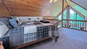 A cozy bedroom with a wooden ceiling, a bed with a quilt, a ceiling fan, and a view of the outdoors through large windows.