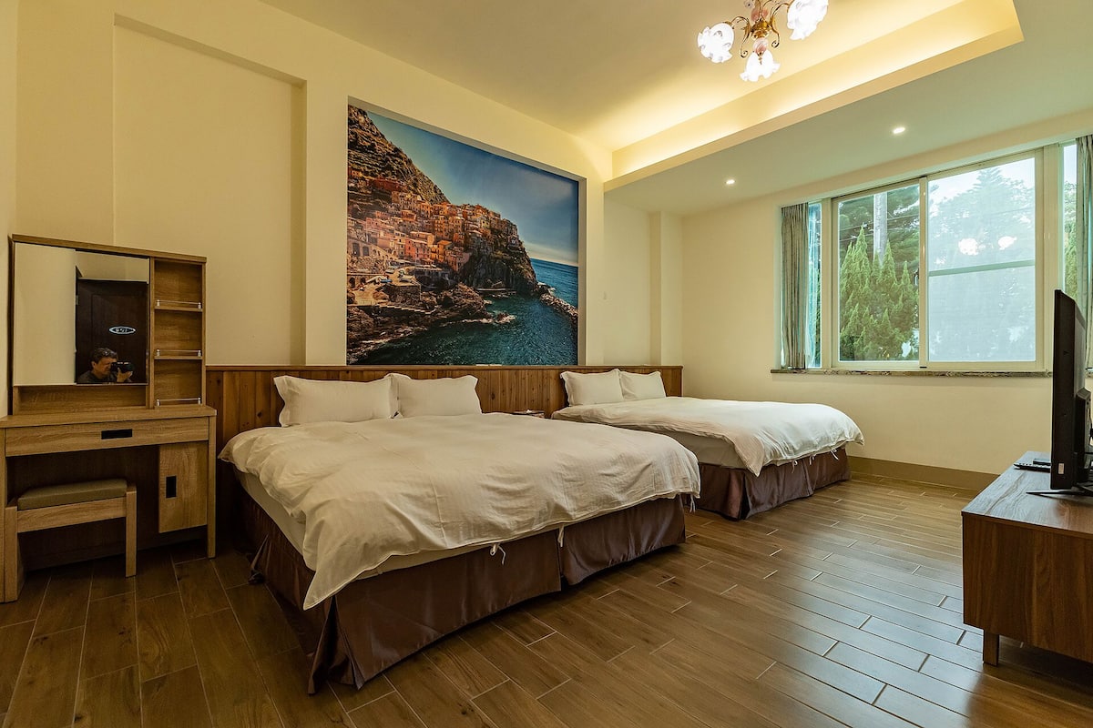 A hotel room with two beds, a wooden desk, a mirror, and a large window.