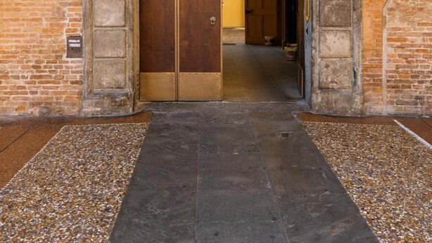 Una puerta de madera grande y arqueada con manija de metal, ubicada en un edificio de ladrillo con piso de baldosas.