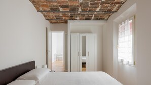 Un dormitorio con techo de ladrillo, paredes blancas, una cama con ropa de cama blanca y una ventana con cortinas translúcidas.