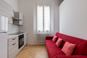 A modern kitchen with white cabinetry, a red sofa, and a window with white curtains. - Appartamento Papavero by Wonderful Italy (Bologna)
