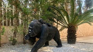 Een beeld van een gorilla in een tuin met kiezelstenen, bomen en een stenen muur.