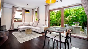 Un salon moderne comprenant un canapé, une table à manger et des chaises, de grandes fenêtres avec des rideaux et une vue sur la verdure à l’extérieur.