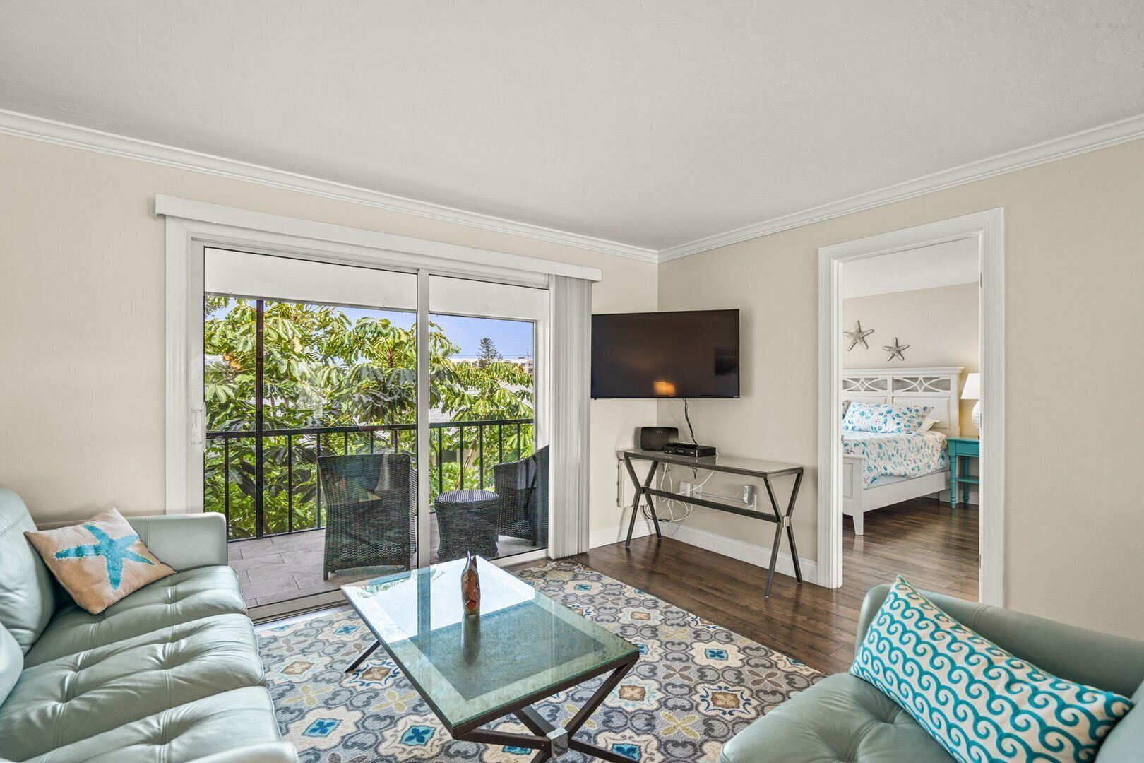 A living room with a glass coffee table, a sofa, a TV mounted on the wall, and a balcony with outdoor furniture.