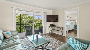 A living room with a glass coffee table, a sofa, a TV mounted on the wall, and a balcony with outdoor furniture. - Pristine Tropical Condo w/ Heated Pool. Walk to Beach, Dining, Shopping & More! VH 304C (Siesta Key)