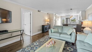 A living room with a sofa, a coffee table, and a TV. There is a kitchen in the background with cabinets and a window. - Pristine Tropical Condo w/ Heated Pool. Walk to Beach, Dining, Shopping & More! VH 304C (Siesta Key)