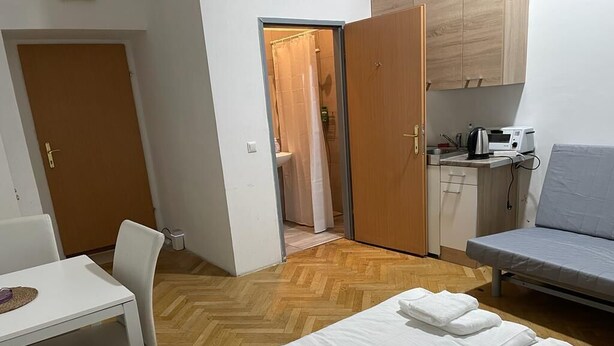 A compact hotel room with a bed, a small table, a chair, a kitchenette, and a shower area.