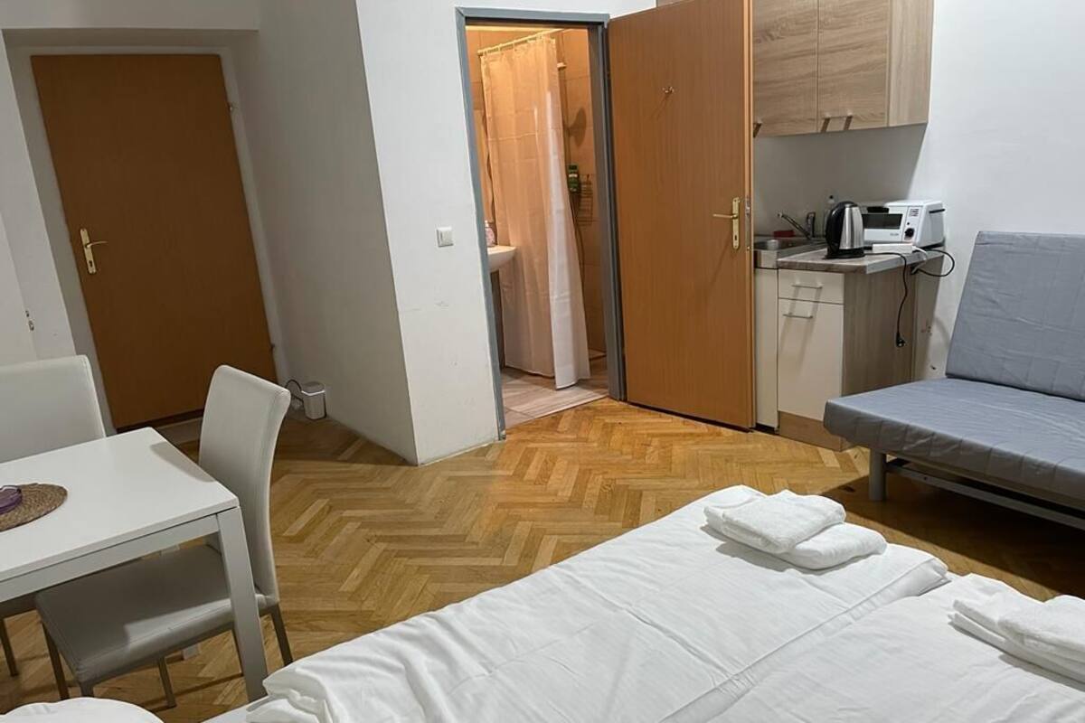 A compact hotel room with a bed, a small table, a chair, a kitchenette, and a shower area.