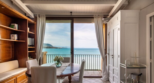 Ein Balkon mit Esstisch, Stühlen und einem Glastisch. Dort steht ein Bücherregal und ein Wagen mit Küchenutensilien. Der Balkon bietet Blick auf ein Gewässer und eine entfernte Küstenlinie.